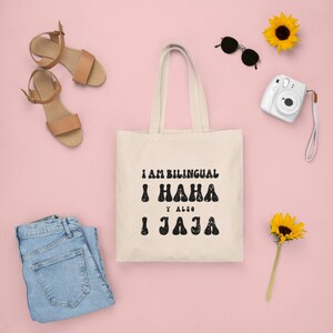 May include: A white canvas tote bag with black text that reads "I am bilingual I haha y also I jaja". The bag is on a pink background with a pair of brown sandals, sunglasses, a camera, and sunflowers.