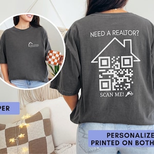 May include: A charcoal gray t-shirt featuring a small logo on the front left chest and a large white graphic on the back. The back design displays a house outline, a QR code, the text "NEED A REALTOR?" and "SCAN ME!" with a small key icon.
