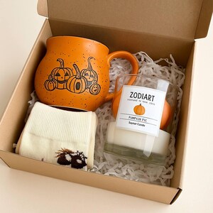 Op de afbeelding: Een herfstcadeaupakket met een oranje mok met pompoengezichten, een kaars en sokken. De kaars zit in een glas met een label met de tekst "Zodiart" en "Pumpkin Pic". De sokken zijn crèmekleurig met een bruin ontwerp.