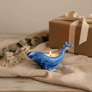 Puede incluir: Un portavelas azul con forma de ballena con una vela encendida en su interior. La ballena de cerámica tiene detalles blancos y está sobre una tela beige. En el fondo hay una caja de regalo envuelta con una cinta crema. Tema decorativo y náutico.