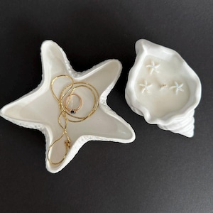 Op de afbeelding: Twee witte keramische sieradenbakjes in de vorm van een zeester en een schelp. Het zeesterbakje bevat een gouden ketting en een ring. Het schelpbakje bevat drie kleine witte zeesterren.