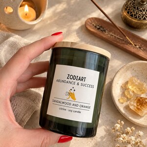 May include: A dark green glass candle jar with a light wood lid and a white label. The label features the text ZODIART, ABUNDANCE & SUCCESS, SANDALWOOD AND ORANGE, and citrine soy candle, along with a graphic of a hand holding a crystal.