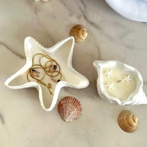 Peut inclure: Un plat en forme d'étoile de mer en céramique blanche avec une chaîne dorée et des anneaux à l'intérieur, un bougeoir en forme de coquillage en céramique blanche avec de la cire blanche et trois coquillages blancs sur le dessus, et quatre coquillages sur une surface en marbre blanc.