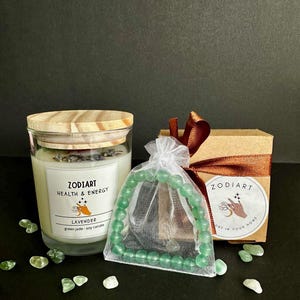 Könnte beinhalten: Ein grünes Jade-Armband und eine Sojawachskerze in einem Glasgefäß mit einem Holzdeckel. Die Kerze ist mit "Zodiart Health & Energy Lavender Green Jade Soy Candle" beschriftet. Das Armband befindet sich in einem weißen Organzasäckchen mit einem braunen Band. Die Kerze und das Armband liegen auf einer schwarzen Oberfläche mit verstreuten grünen Jadesteinen.