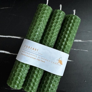 Puede incluir: Tres velas verdes con patrón de panal de abeja agrupadas. Una etiqueta blanca alrededor de las velas dice "ZODIART" y "ABUNDANCE & MONEY CANDLE" con las notas de fragancia "sándalo y naranja, salvia".