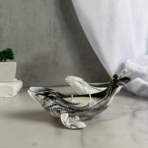 Può includere: Una candela a forma di balena bianca e nera con un designo marmoreo. La candela ha due stoppini ed è fatta di cera di soia.