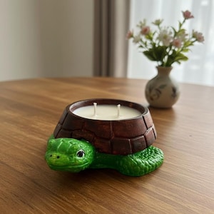 Puede incluir: Un portavelas con forma de tortuga con caparazón marrón oscuro y cabeza y patas verdes. El portavelas contiene una vela blanca con dos mechas. Está sobre una mesa de madera.