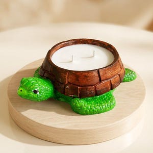 Puede incluir: Un portavelas con forma de tortuga con caparazón marrón y cuerpo verde. El portavelas está sobre una base redonda de madera. La vela interior es blanca y tiene dos mechas.