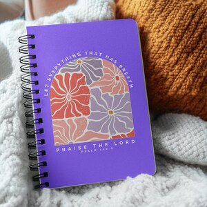 May include: A purple spiral-bound notebook with the text "LET EVERYTHING THAT HAS BREATH PRAISE THE LORD" and Psalm 150:6. The cover features a floral design in shades of red, pink, and purple. The notebook rests on a white knitted blanket.