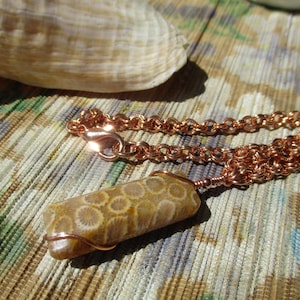 May include: A copper chain necklace with a fossil pendant. The pendant is a light brown color with a circular pattern. The necklace is displayed on a patterned surface with a seashell in the background.