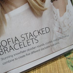 May include: A product display card for Sofia Stacked Bracelets featuring black text on a white background. The text reads: SOFIA STACKED BRACELETS. Stunning snowflake obsidian rounds, crystals and gold findings combine to create five different stackable bracelets.