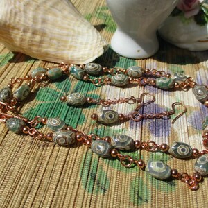 May include: A handmade jewelry set featuring a necklace and earrings. The necklace and earrings are made with patterned beads in shades of green and gray, strung on copper chains. The set is displayed on a patterned surface with a seashell and decorative items.