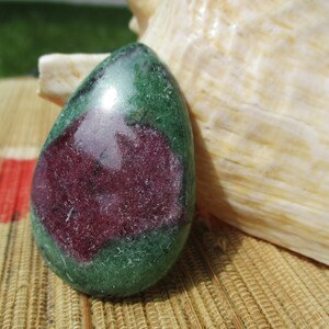 May include: A polished, teardrop-shaped ruby zoisite gemstone featuring a deep magenta-red center surrounded by a mottled forest green and black mineral matrix, displaying a smooth, reflective surface with natural color variations throughout.