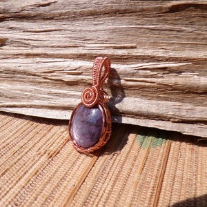 May include: A handcrafted copper wire pendant with a purple oval stone. The wire is intricately woven around the stone and features a spiral design at the top. The pendant is displayed on a textured wooden surface.