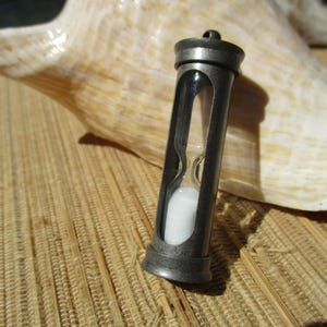May include: A small hourglass pendant featuring a dark metallic frame with a cylindrical glass center containing white sand. The top of the frame includes a small metal loop for attachment, and the design is vertical with a clear view of the sand.