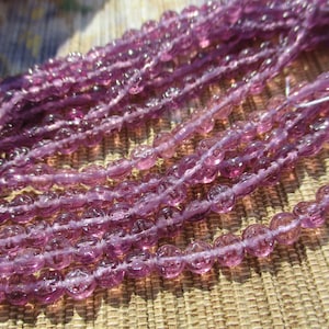 May include: Strands of translucent, light purple beads. Each bead is round with a textured surface. The beads are strung together, creating a flowing, organic pattern. The beads are a soft, muted purple color, and the image is well-lit.