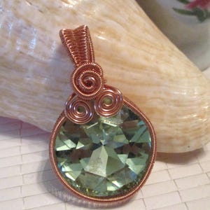 May include: A handcrafted pendant featuring a faceted, round, light green gemstone. The stone is encased in copper wire with decorative spiral accents and a bail for hanging. The pendant is displayed against a neutral background.