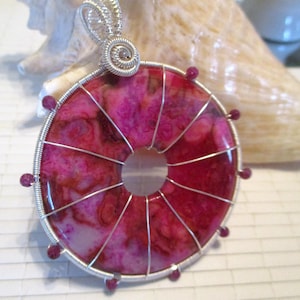 May include: A handcrafted pendant featuring a vibrant pink and red agate stone with a circular design. The stone is encased in a silver wire frame with a spiral bail and small red beads. The pendant is a unique piece of jewelry.