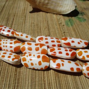 May include: A strand of oblong beads featuring a white base with irregular orange spots. The beads are arranged in a multi-row formation, showcasing a smooth, polished finish and a consistent pattern across each individual piece in the collection.