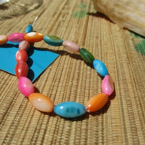 May include: A strand of oval-shaped beads in a variety of colors including bright pink, orange, light blue, green, and coral. The beads have a smooth, polished finish and are strung together on a thin wire, partially resting on a blue card.