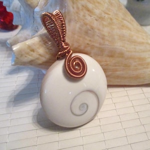 May include: A handcrafted pendant featuring a white, oval-shaped stone with a spiral design. The stone is adorned with copper wire wrapping, including a decorative spiral at the top. The pendant is a unique piece of jewelry.