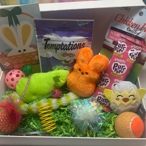 May include: A white gift box filled with Easter-themed pet toys and treats. The box contains a green plush mouse, an orange plush bunny, a white and silver plush chick, a pink and white ball, a spring toy, a sparkly toy, a tennis ball, and several bags of pet treats. The box is decorated with green grass and a card with a bunny on it.