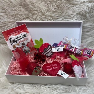 May include: A white gift box filled with pink and red tissue paper. The box contains a variety of cat treats, toys, and a heart-shaped pillow that says "Strawberry Much Love".