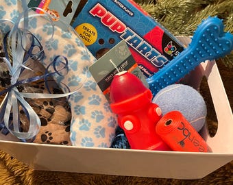 May include: A white gift basket filled with dog toys and treats. The basket includes a blue and white patterned dog bed, a blue and white chew toy, a red fire hydrant chew toy, a blue and white rope toy, a blue and white tennis ball, a bag of peanut butter flavored dog treats, and a blue and white dog toy with a bone shape.