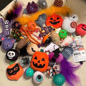 May include: A collection of Halloween-themed cat toys, including plush pumpkins, ghosts, bats, and skeleton figures. The toys are in various colors, such as orange, purple, black, and white. Some toys have feathers and dangling strings.