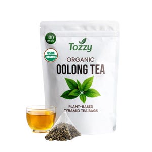 May include: A white bag of Tozzy Organic Oolong Tea with a green USDA Organic seal and the text "100 BAGS". A glass of tea and a pyramid tea bag are in front of the bag. The bag has the text "PLANT-BASED PYRAMID TEA BAGS".