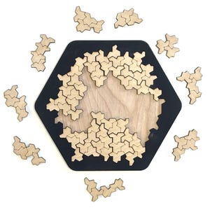 May include: A wooden puzzle with a black hexagonal frame and a light wood interior. The puzzle is partially assembled, with several small, interlocking pieces scattered around the frame. The pieces are a natural wood color, and the overall design is geometric.