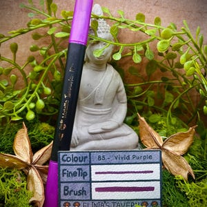Könnte beinhalten: Ein leuchtend violetter Fein- und Pinselstift mit dem Text "Colour 85 - Vivid Purple" und "ELINIA'S TAVER" auf einem kleinen Schild. Der Stift befindet sich vor einem Hintergrund aus Grün und einer kleinen Buddha-Statue.