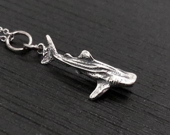 Puede incluir: Un collar de plata con un colgante detallado de tiburón ballena. El colgante cuelga de una cadena de plata.