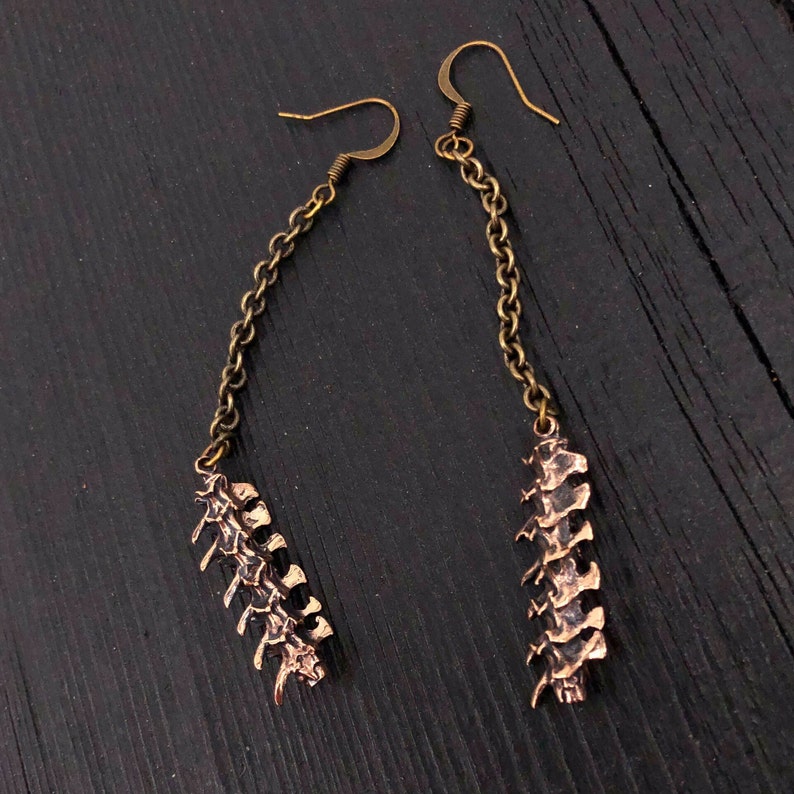 May include: A pair of gold-toned earrings with a dangling spine-shaped charm. The earrings are made of metal and have a delicate chain.