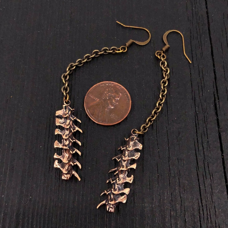 May include: A pair of long, dangling earrings with a bronze chain and a bronze pendant shaped like a spine. The earrings are hanging from a penny for scale.