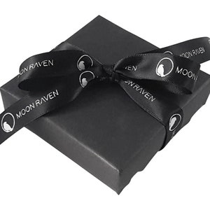 May include: A black gift box with a black ribbon tied in a bow. The ribbon has the words "Moon Raven" printed on it in white.