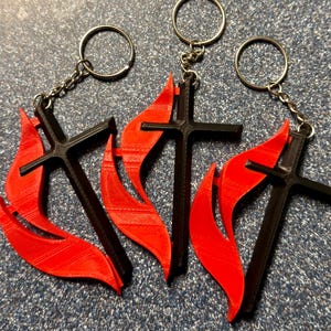May include: Three keychains featuring a black cross design with a red flame accent. Each keychain has a silver-colored ring and chain. The keychains are displayed on a textured, dark blue surface.