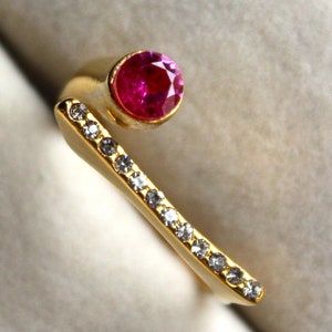 May include: A gold ring with a single pink gemstone and a row of smaller clear gemstones.