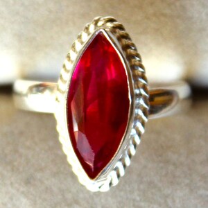 Könnte beinhalten: Ein silberner Ring mit einem marquiseförmigen, tiefroten Edelstein. Der Edelstein ist in einer silbernen Lünette mit einem gedrehten Seildesign gefasst. Der Ring hat ein glattes, poliertes Band, das einen Kontrast zur strukturierten Lünette bildet.