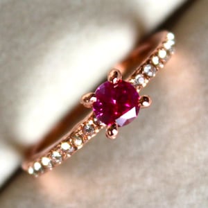 Pode incluir: Anel de ouro rosa com uma vibrante gema de rubi redonda no centro. A banda é adornada com pequenas pedras brilhantes. O design do anel é delicado e elegante, adequado para várias ocasiões.