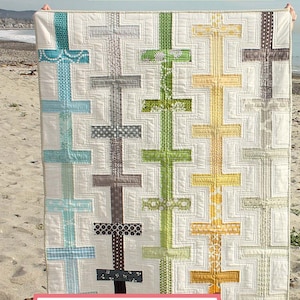 Könnte beinhalten: Eine weiße Steppdecke mit einem Muster aus bunten Kreuzen. Die Steppdecke wird an einem Sandstrand ausgestellt, mit dem Text "Crossroads" in einem rosa Banner am unteren Rand des Bildes. Der Text "Amy Smart Brilliant Patterns" befindet sich in einem weißen Banner am oberen Rand des Bildes.