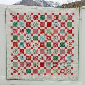 May include: A red and green Christmas quilt with a checkered pattern. The quilt is 60 inches by 60 inches and features a variety of festive prints. The quilt is hanging on a white fence with a snowy mountain in the background. The text "pattern by Amy Smart" is at the bottom of the image.