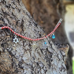 May include: A bracelet featuring a red and silver chain, adorned with charms such as a Star of David, a Hamsa hand, and an evil eye. The bracelet is set against a textured tree trunk, with a blurred green backdrop.