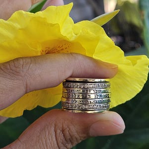 May include: A gold-toned ring with multiple bands, each inscribed with text and adorned with small, clear stones. The ring is held in front of a bright yellow flower, with green foliage in the background.