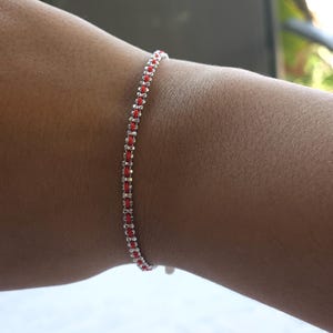 Könnte beinhalten: Ein zartes Armband mit einer Reihe kleiner, silberfarbener Glieder, die durch leuchtend rote Fäden verbunden sind. Das Armband ist für das Handgelenk konzipiert und zeigt eine schlichte, aber elegante Ästhetik. Das Armband ist etwa 18 cm lang.