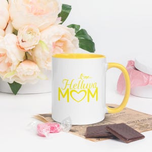 May include: White ceramic mug with a yellow rim and handle. The mug has a yellow graphic that says "One Helluva Mom" with a heart in the middle of the word "Mom".