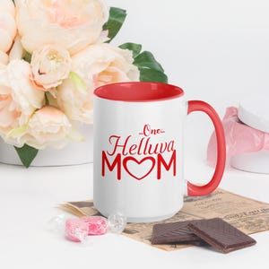 May include: White ceramic coffee mug with a red interior and handle. The mug has a red graphic that says "...One...Helluva MOM" with a red heart in the middle of the word MOM.