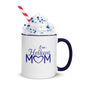 May include: White ceramic mug with a blue rim and handle. The mug has a blue design that says "...One...Helluva MOM" with a heart in the middle of the word MOM. The mug has whipped cream with blue sprinkles on top and a red and white striped straw.