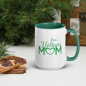Puede incluir: Taza de café de cerámica blanca con borde y asa verdes. La taza tiene un diseño verde que dice "...One...Helluva MOM" con un corazón verde.