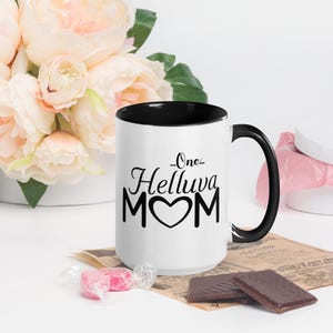 May include: White ceramic coffee mug with a black rim and handle. The mug has a black graphic that says "...One...Helluva MOM" with a black heart in the middle of the word MOM.